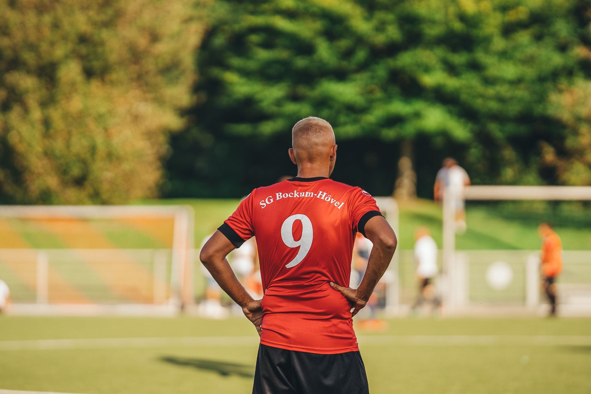 Back view of a soccer player in a red jersey on a green field during a game.