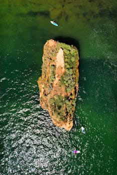 Drone view of paddleboarders circling a rocky island in clear green waters.
