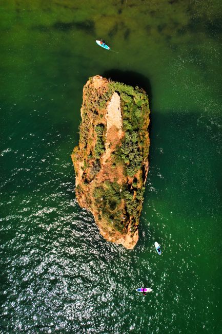 Drone view of paddleboarders circling a rocky island in clear green waters.