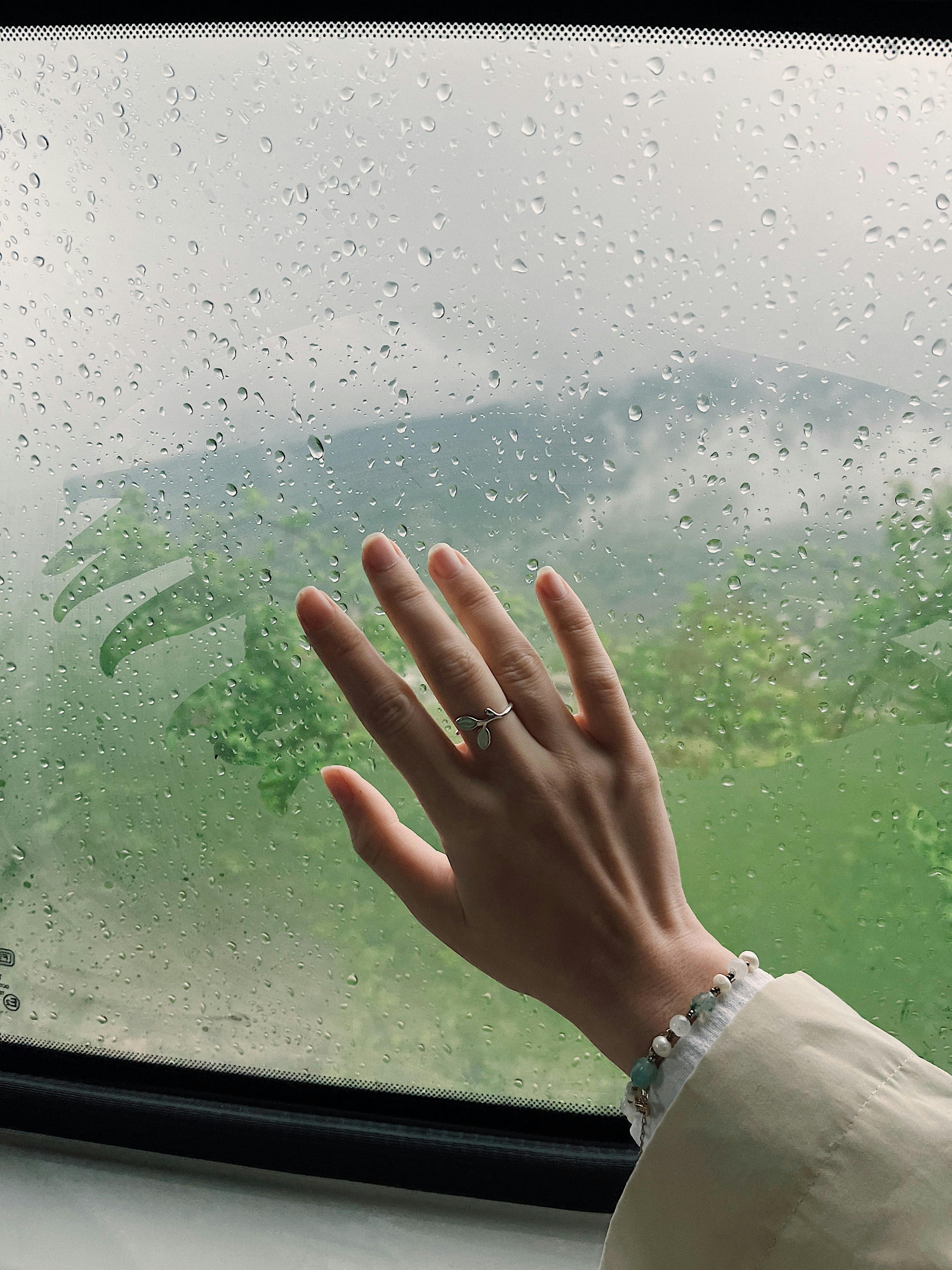 A hand wearing a ring touches a raindrop-covered window overlooking lush greenery.