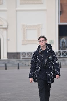 A stylish man in a black patterned coat walks confidently with a camera.