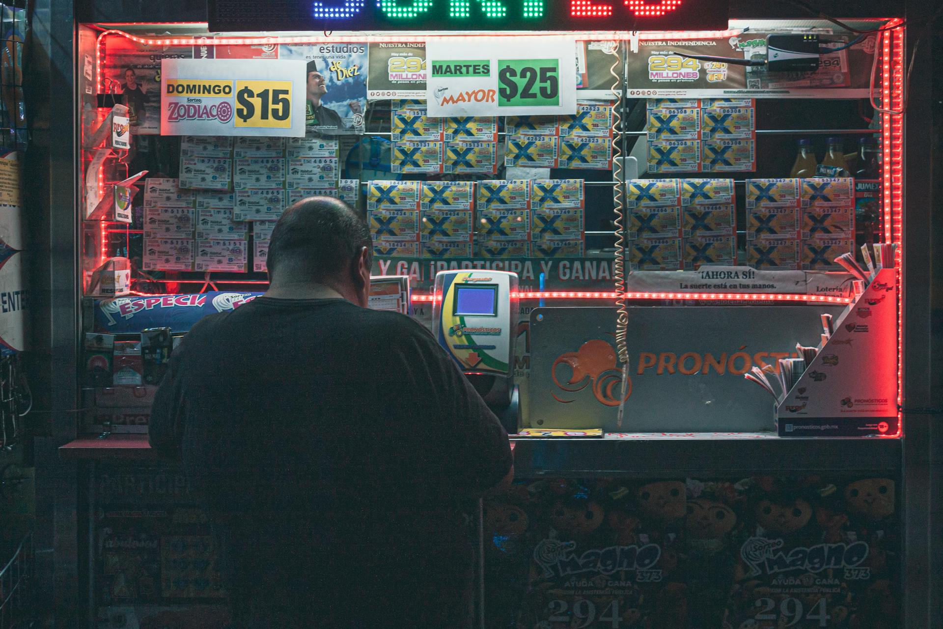 A man purchasing a lottery ticket at an illuminated kiosk in Mexico City, highlighting risk and hope.