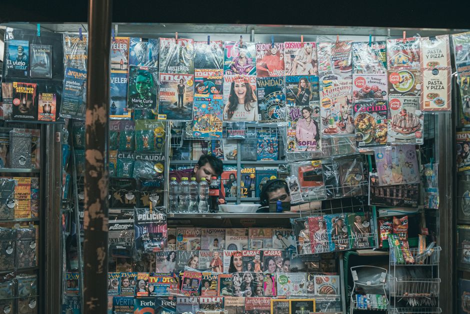 メキシコシティのカラフルな雑誌スタンドには雑誌やスナックが満載で、中に2人の男性が見えます。
