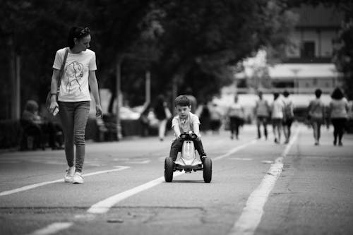 Grayscale Photo of Boy Riding on Toy Car on the Road Beside a Woman