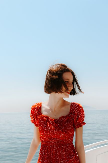 A young woman in a red dress stands on a boat with the ocean and clear blue sky in the background.