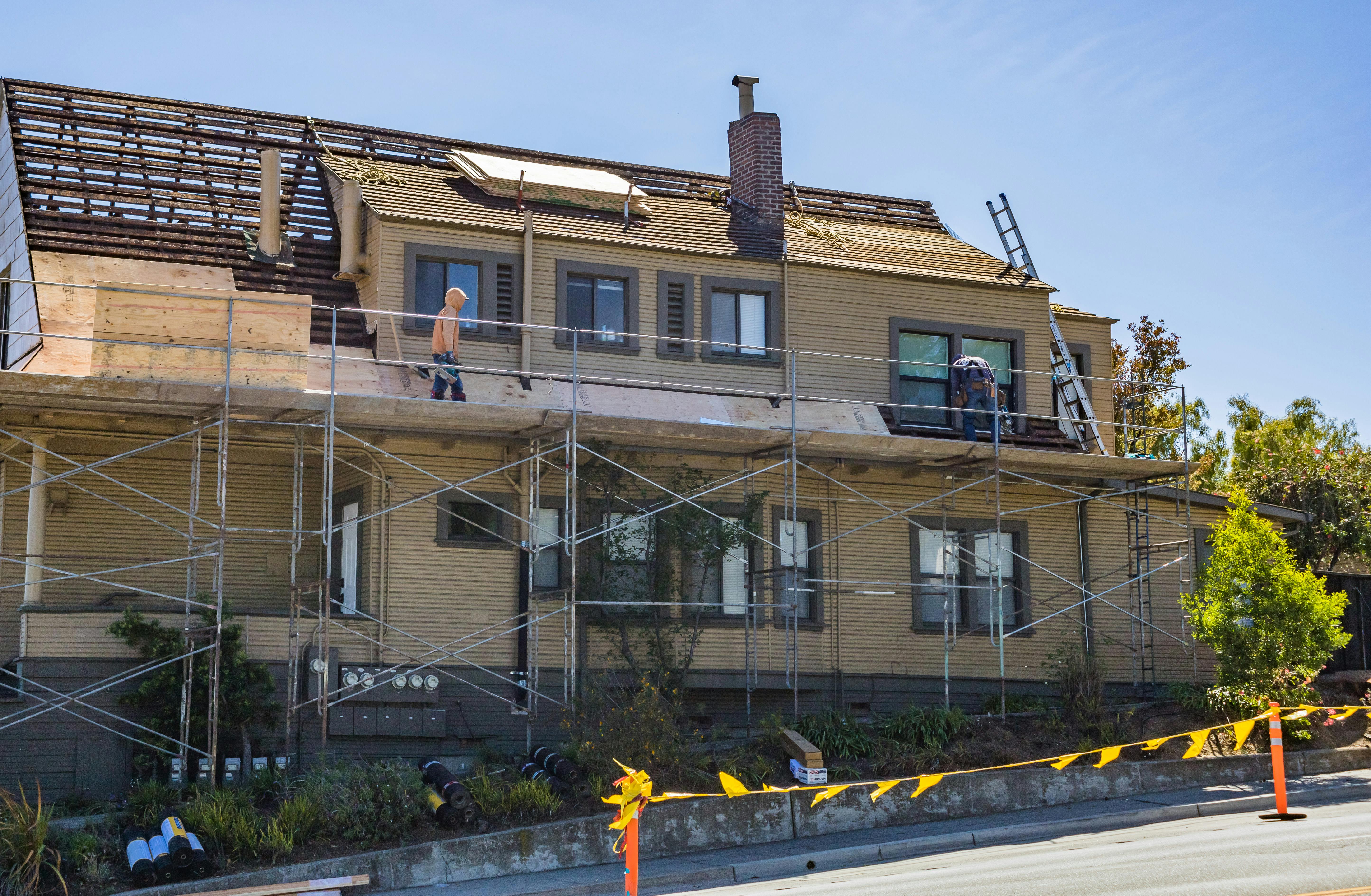 House under renovation with scaffolding and workers fixing the roof on a sunny day.