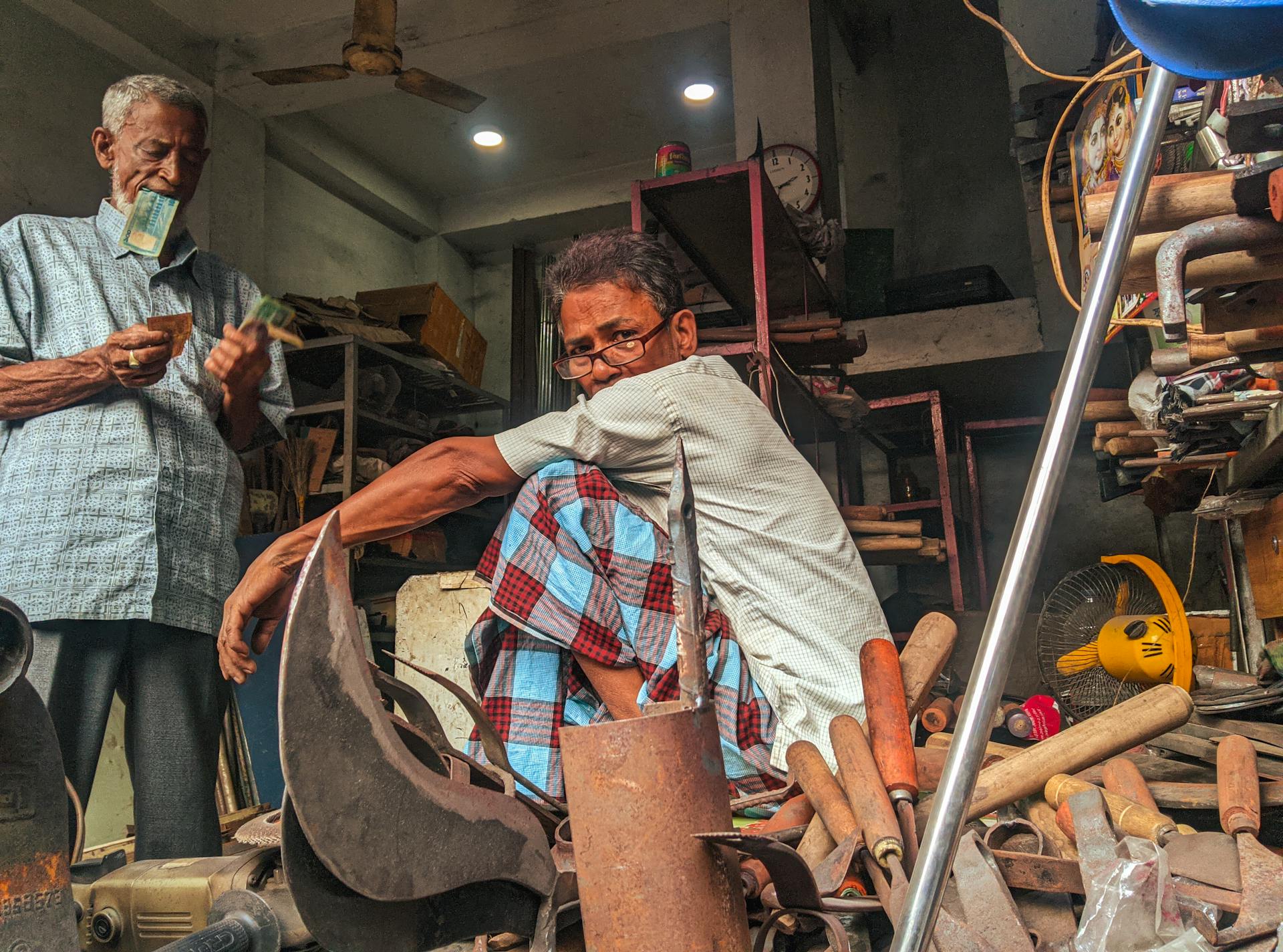 Two elderly men in a busy Bangladeshi workshop exchanging money surrounded by tools and products.