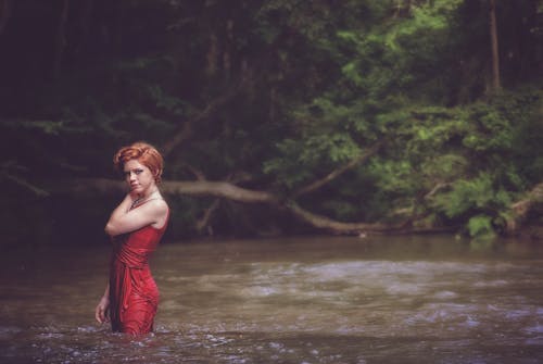 森の近くの水域に赤いひもスパゲッティストラップドレスの女性