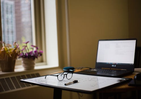 A serene indoor workspace featuring a laptop, eyeglasses, and a whiteboard near a window with flowers.