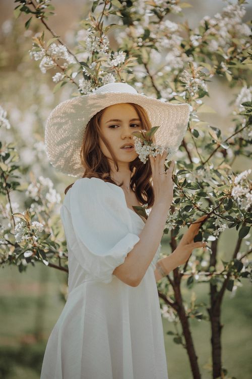 Free Young woman in a white dress and sun hat enjoying a blooming garden in spring. Stock Photo