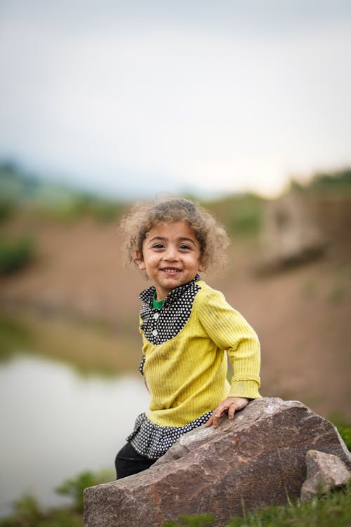 Free A Young Girl in Yellow Sweater Smiling while Standing Near the Rock Stock Photo