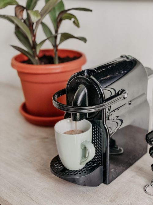 Free A modern coffee machine pours espresso into a white cup beside a potted plant, creating a cozy kitchen scene. Stock Photo