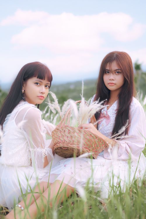 Free Beautiful portrait of two women in white dresses sitting in a grassy field with a basket. Stock Photo
