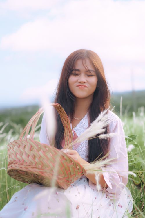 Free Cute young woman holding a woven basket while sitting in a grassy field outdoors. Stock Photo
