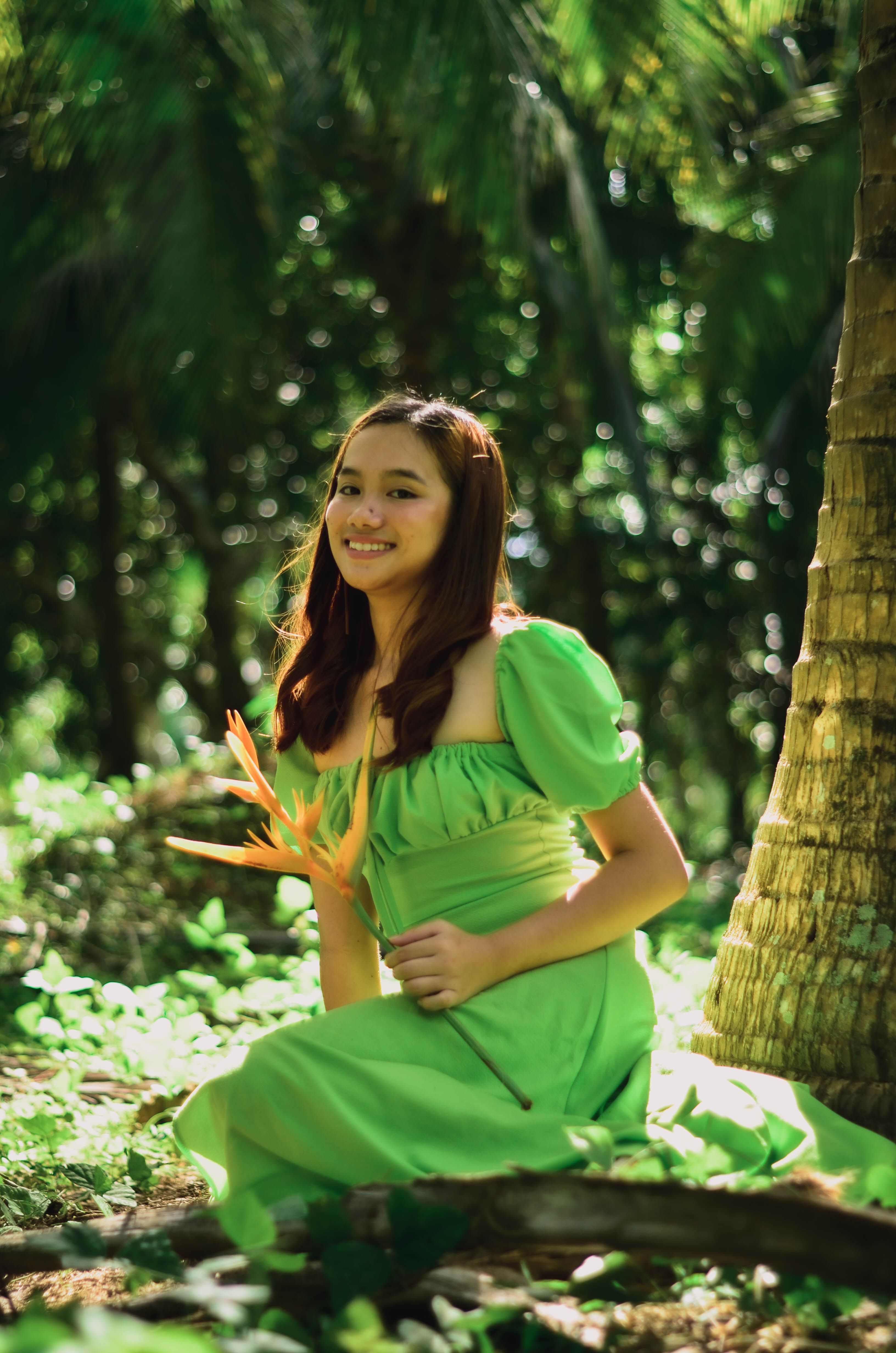 A young woman in a green dress smiles while holding a tropical flower in an outdoor setting.
