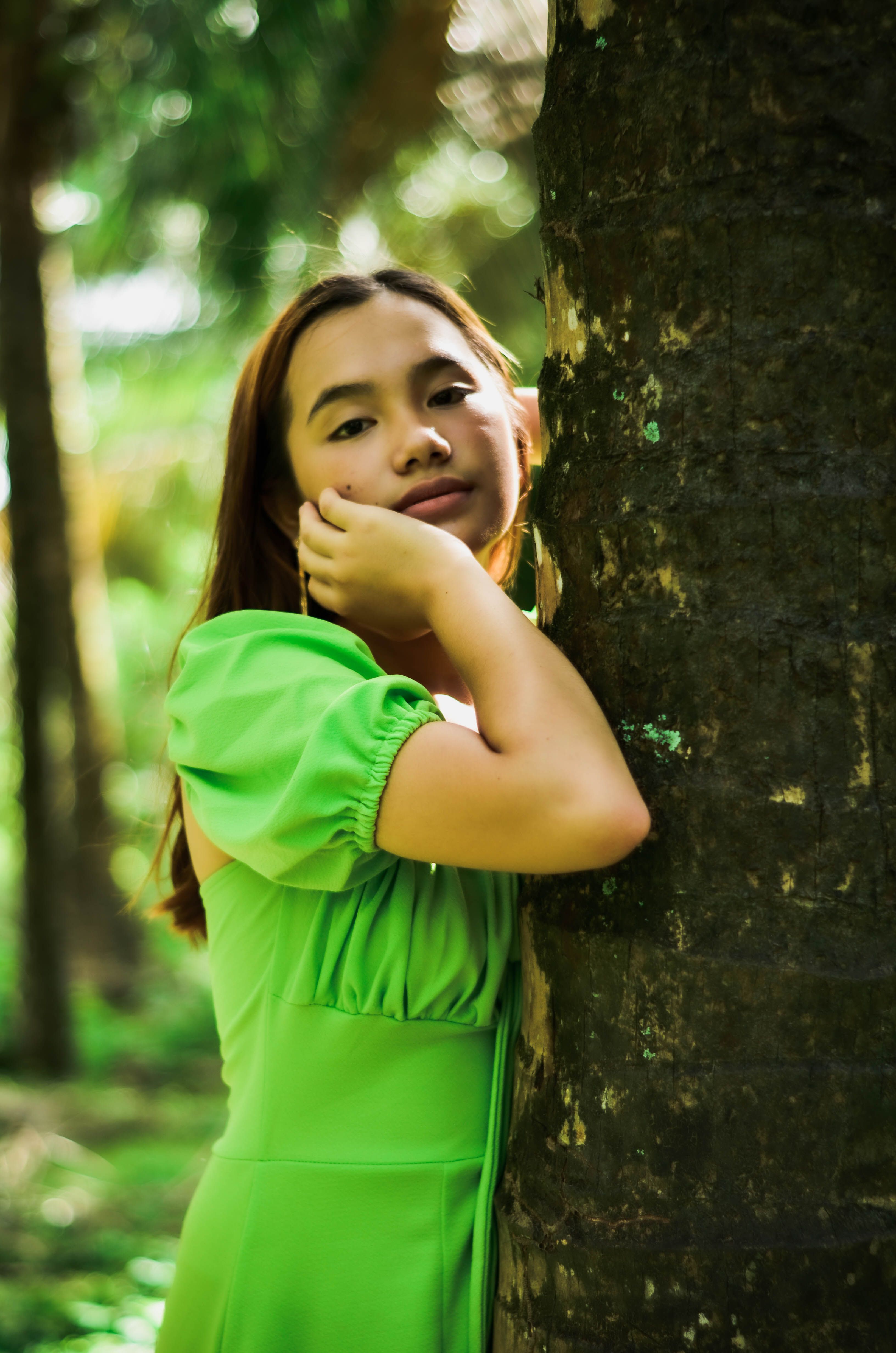 Portrait of a young woman in a green dress leaning against a tree trunk in a lush outdoor setting.