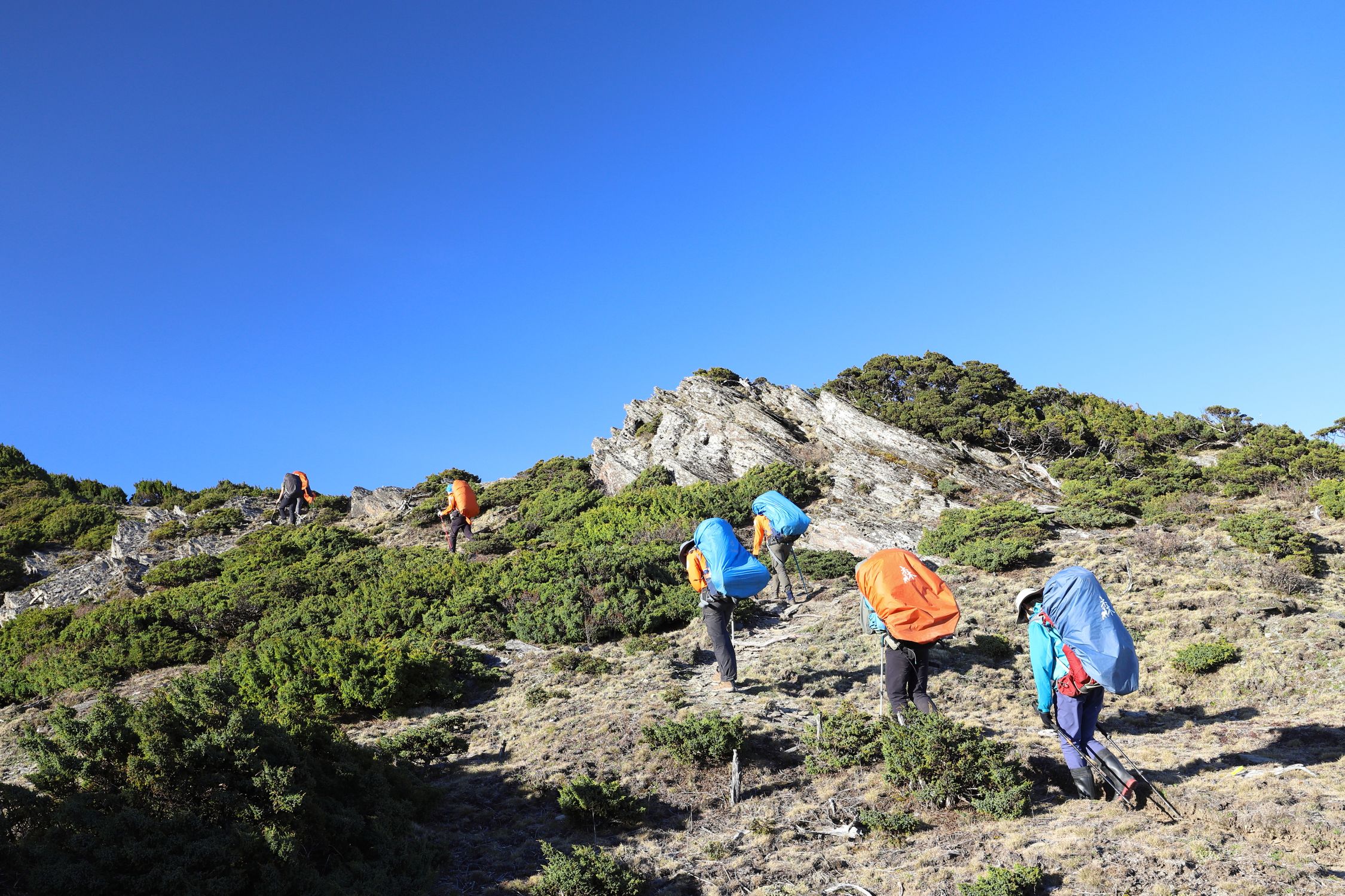 Photo by P Hsuan Wang Hikers with backpacks trekking up a mountainous path under a clear blue sky