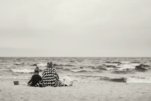 A grayscale photo of a mother and child at the seashore, by the ocean.