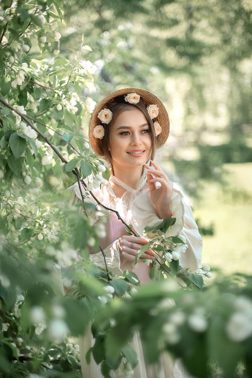 Free A cheerful woman in a floral hat and white long sleeves stands amidst blossoming trees, enjoying a sunny day. Stock Photo