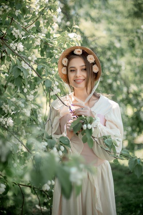 Free Smiling woman in vintage dress and hat surrounded by white blooming flowers outdoors. Stock Photo