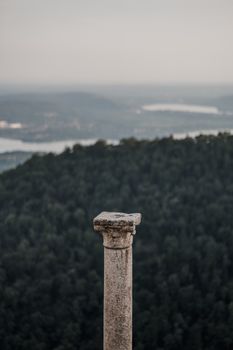A solitary ancient column stands tall with a scenic view of a vast forest and distant river.