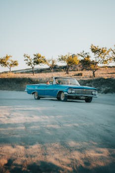 A classic blue convertible on a scenic dirt road with trees under a blue sky.