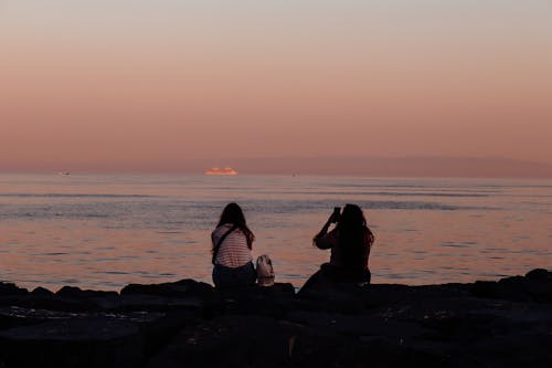 Silhouette of Women Sitting on the Shore at Sunset 