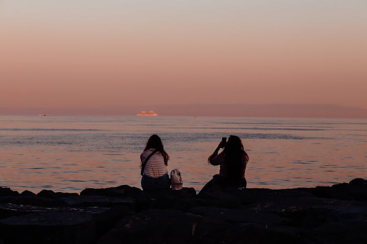 Silhouette Of Women Sitting On The Shore At Sunset 