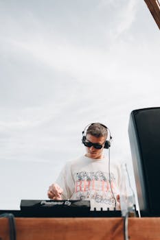 DJ wearing sunglasses and headphones mixes music outdoors under a bright sky for a lively event.