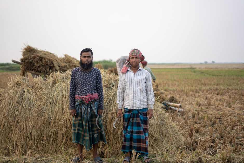 Two farmers in traditional attire standing by haystacks in rural Bangladesh field.