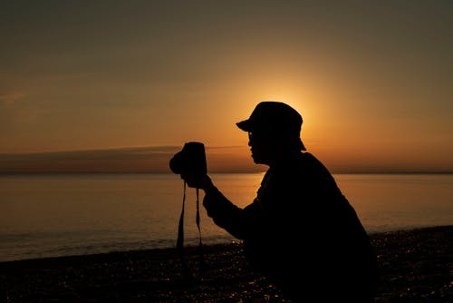 Silhouette of Person Wearing Bucket Hat Standing on the Beach while Taking Photos of the Scenery Using Camera