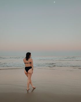 A woman stands on a peaceful beach at sunset, gazing at the horizon in a black bikini.