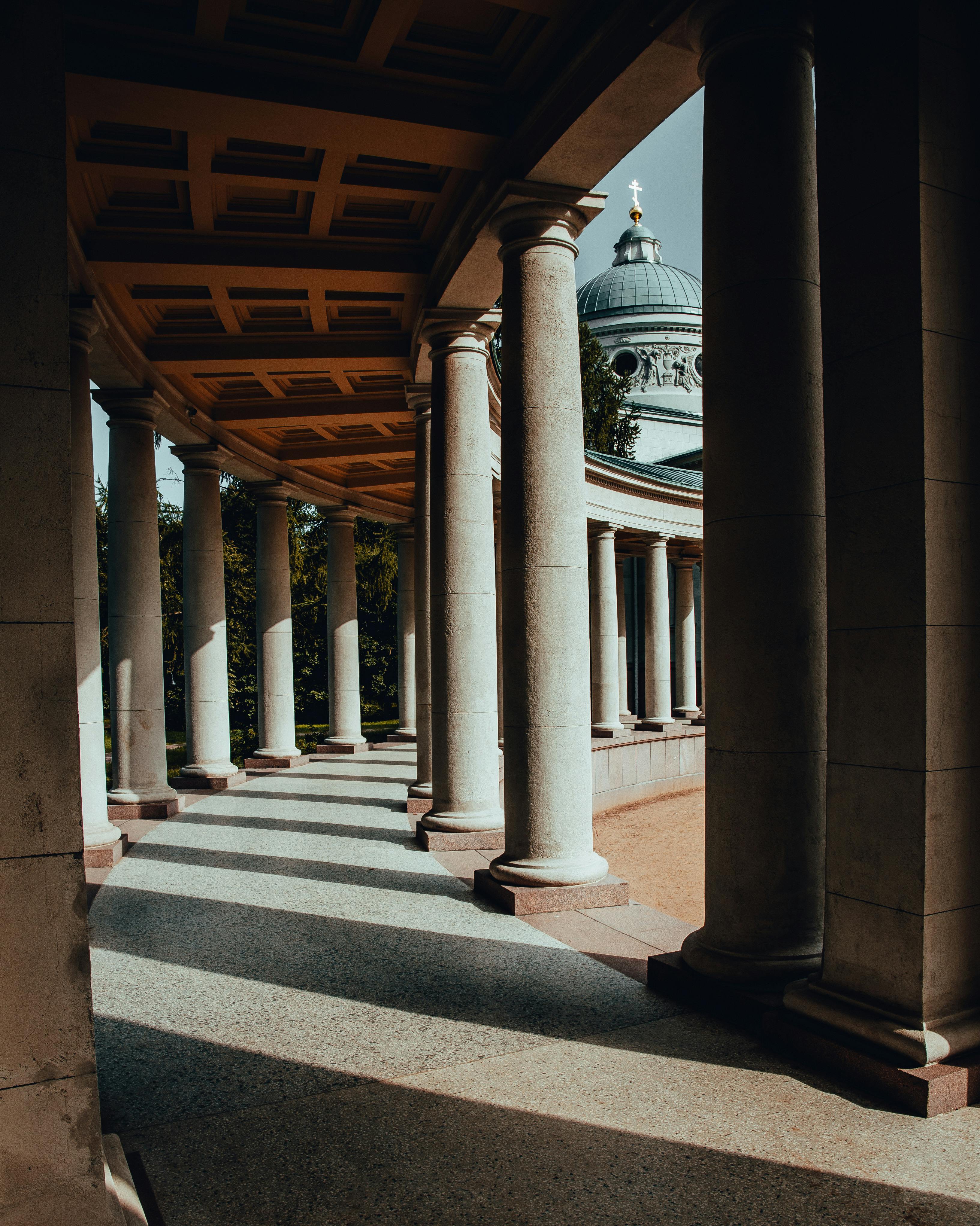 Sunlit pathway with elegant neoclassical columns and a dome in the background, showcasing classical architecture.