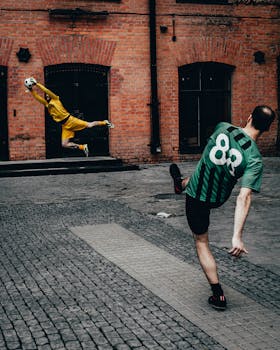 Two male soccer players competing energetically in a street setting against a brick wall backdrop.
