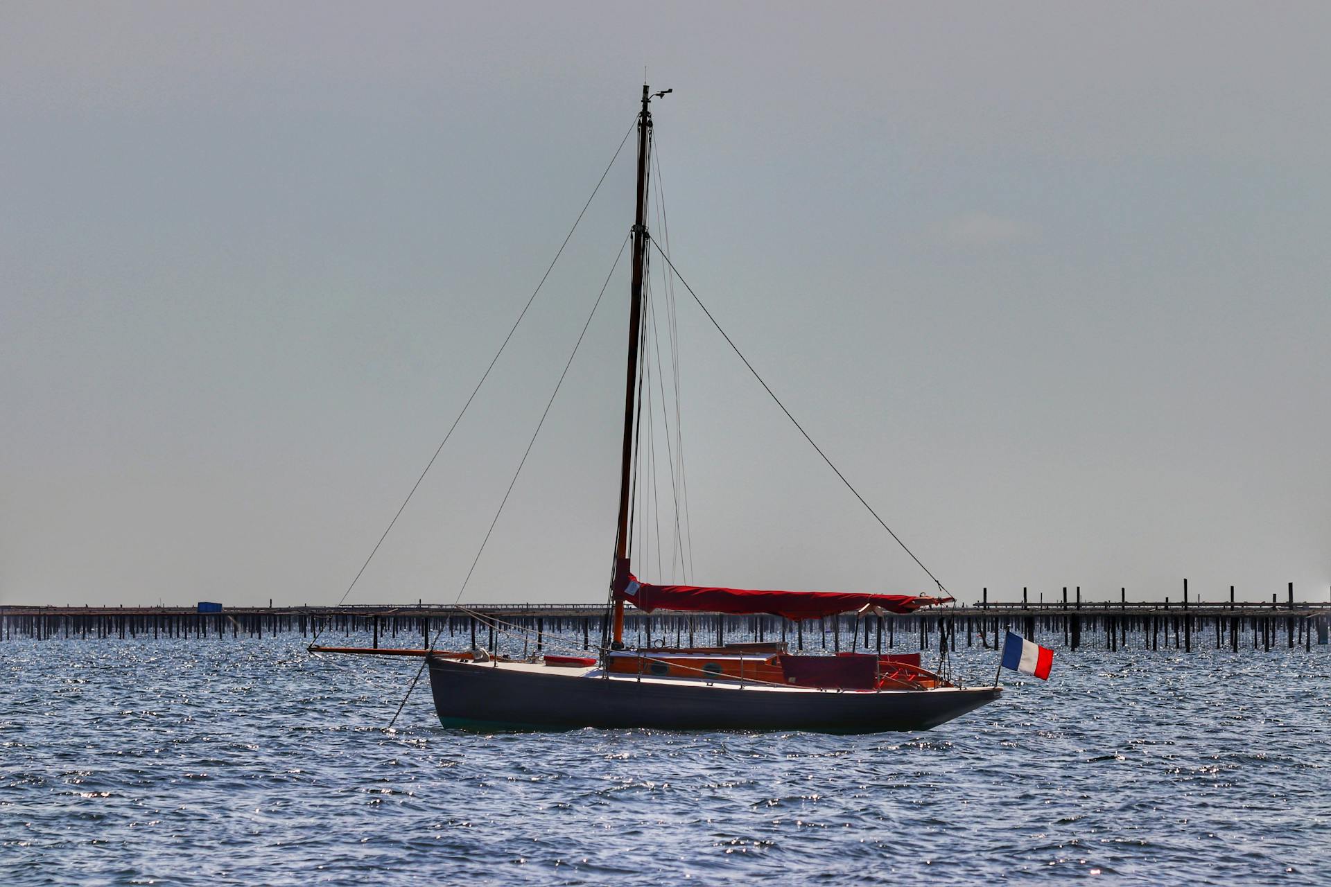 A sailboat displaying the French flag sails on calm waters under an overcast sky.