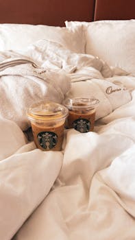 Two Starbucks iced coffees on a cozy bed, wrapped in plush linens for a relaxing morning vibe.
