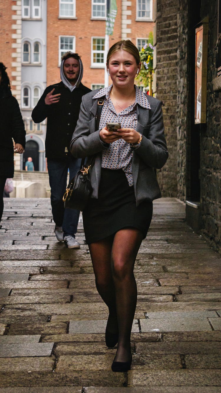 Woman In Gray Blazer And Black Skirt Walking While Holding A Phone 