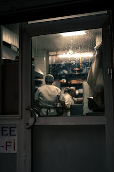 View from outside a kitchen with chefs working, visible through a glass door with a vintage look.