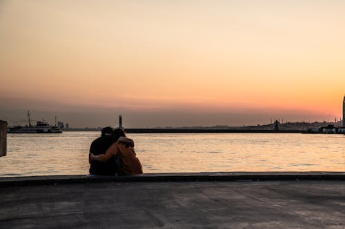 Back View of a Couple Sitting on a Shore at Sunset and Hugging 