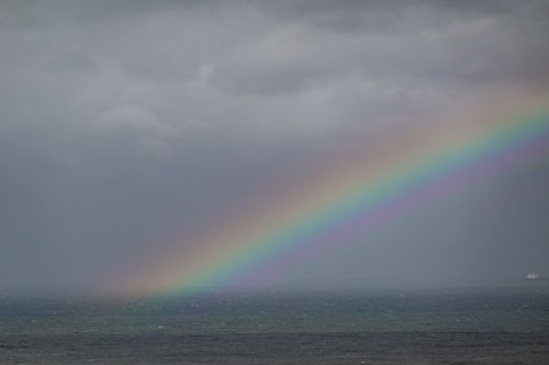 Free A vivid rainbow arcs across a gloomy sky over a calm ocean, creating a scenic view. Stock Photo