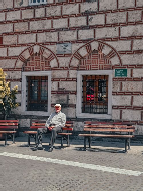 Free Senior man sitting on a bench against a historic brick wall in a sunny urban setting. Stock Photo