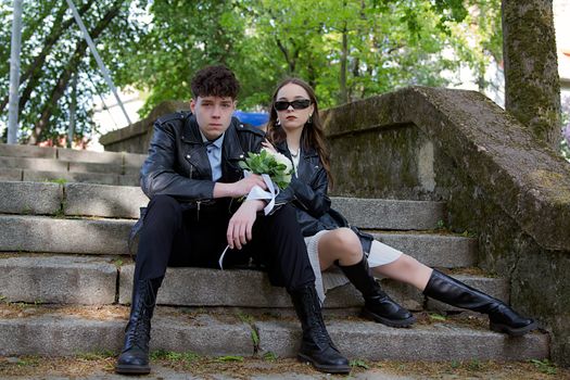 Young couple in leather jackets sitting on outdoor steps holding flowers, showcasing modern style.