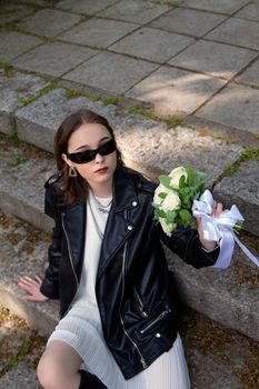 Stylish woman in leather jacket holding flowers, sitting on stone steps outdoors.