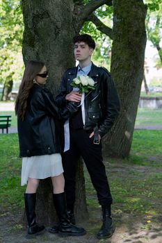 A young couple standing by a tree, wearing leather jackets and holding white roses in a park setting.