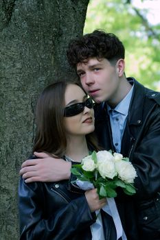 Young couple in leather jackets embracing by a tree with white roses, symbolizing love.