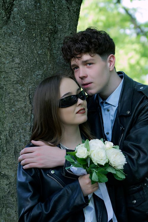 Free Young couple in leather jackets embracing by a tree with white roses, symbolizing love. Stock Photo