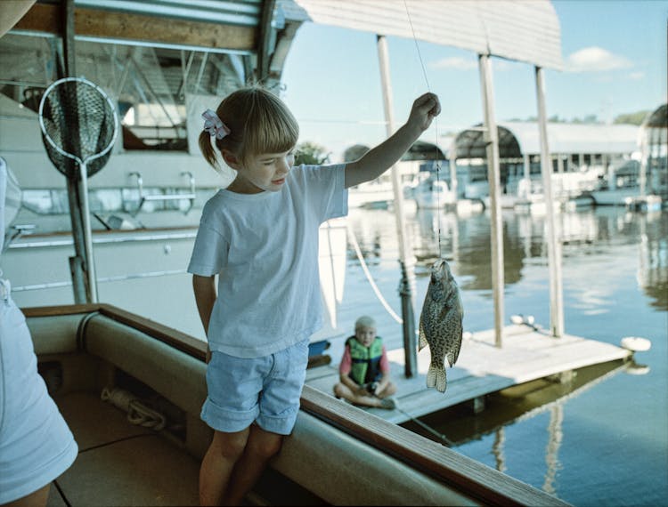 A Little Girl Standing On A Boat And Holding A Fish Caught On A Hook 