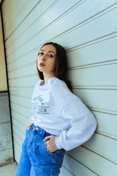 Fashionable young woman leaning against a wall in casual jeans and a white sweatshirt.