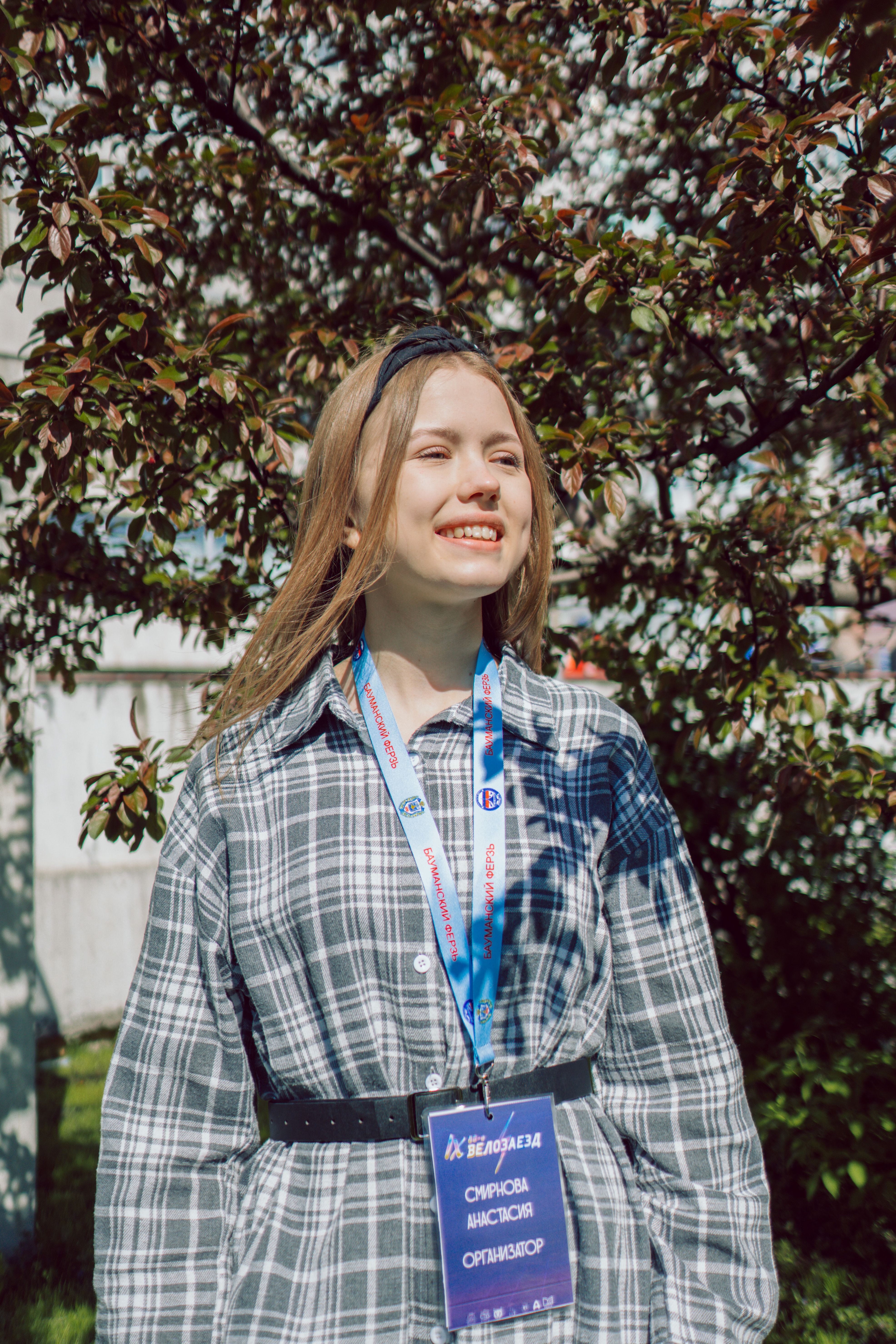 A young blonde woman in plaid is smiling under a sunny sky, standing outdoors with a badge around her neck.