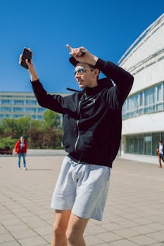 Man in black jacket and sunglasses raising arms joyfully outdoors.